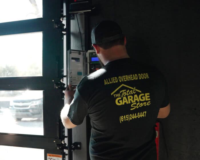 A person in a t-shirt labeled "The Total Garage Store" working on a garage door mechanism.