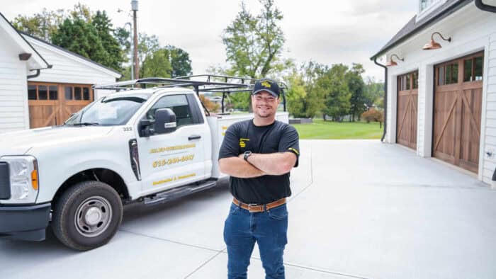 Man in a black shirt and jeans stands with arms crossed in front of a white service truck on a driveway, with houses and trees in the background.