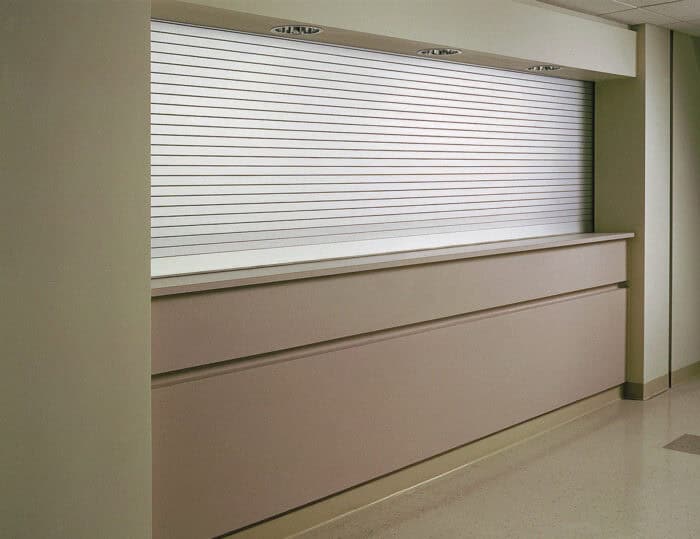 Closed beige reception desk with a metal roll-up shutter. Vertical panels cover the desk's front, and there are overhead lights on the ceiling. Floor is light-colored.