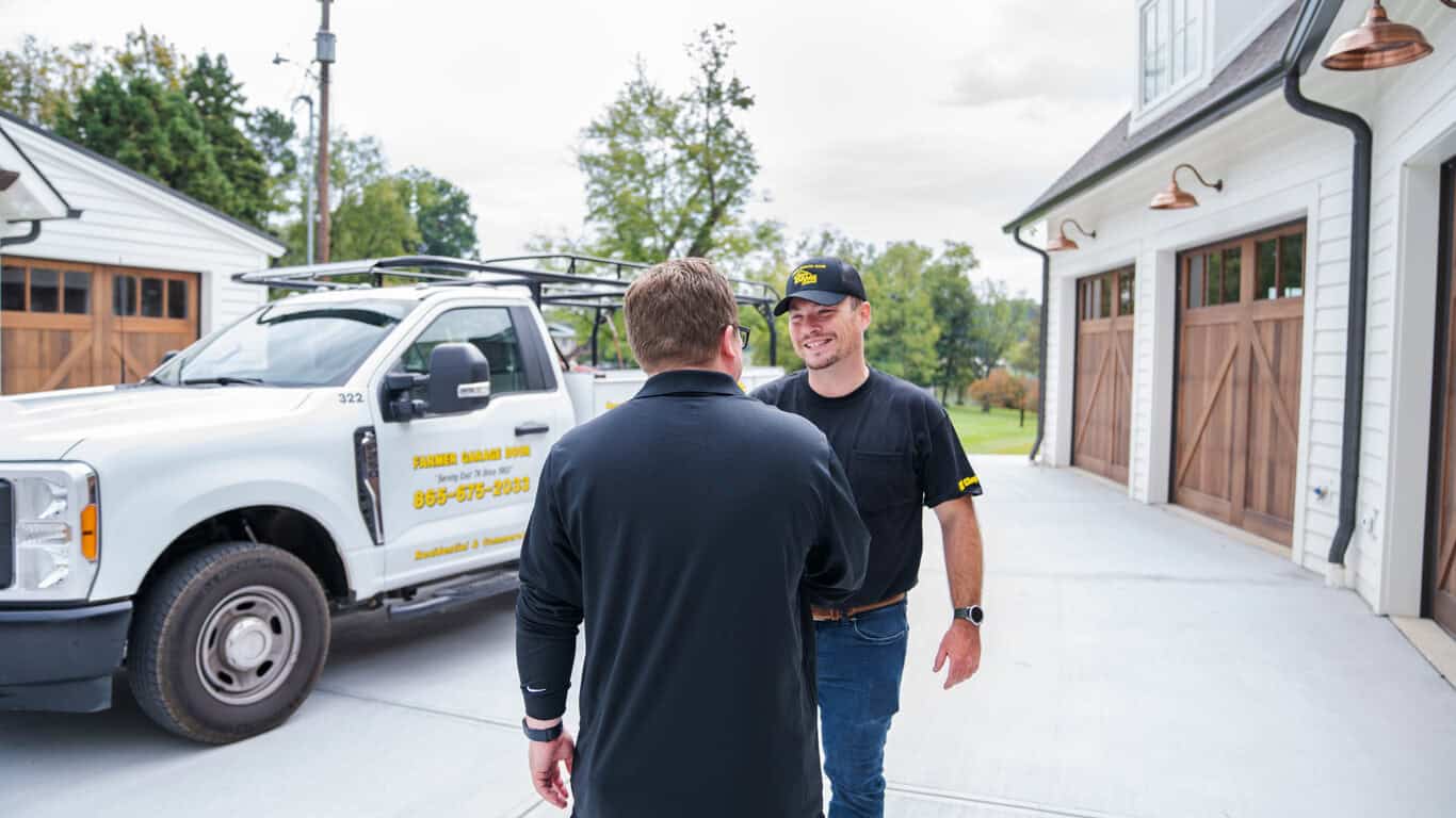 Two men are shaking hands in a driveway. A pickup truck with company signage is parked nearby. In the background, a building features large wooden residential garage doors, highlighting their recent garage door replacement project.
