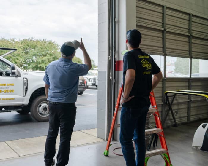 Two men stand in a garage next to a ladder, with one pointing outside. A truck is partially visible near the entrance.