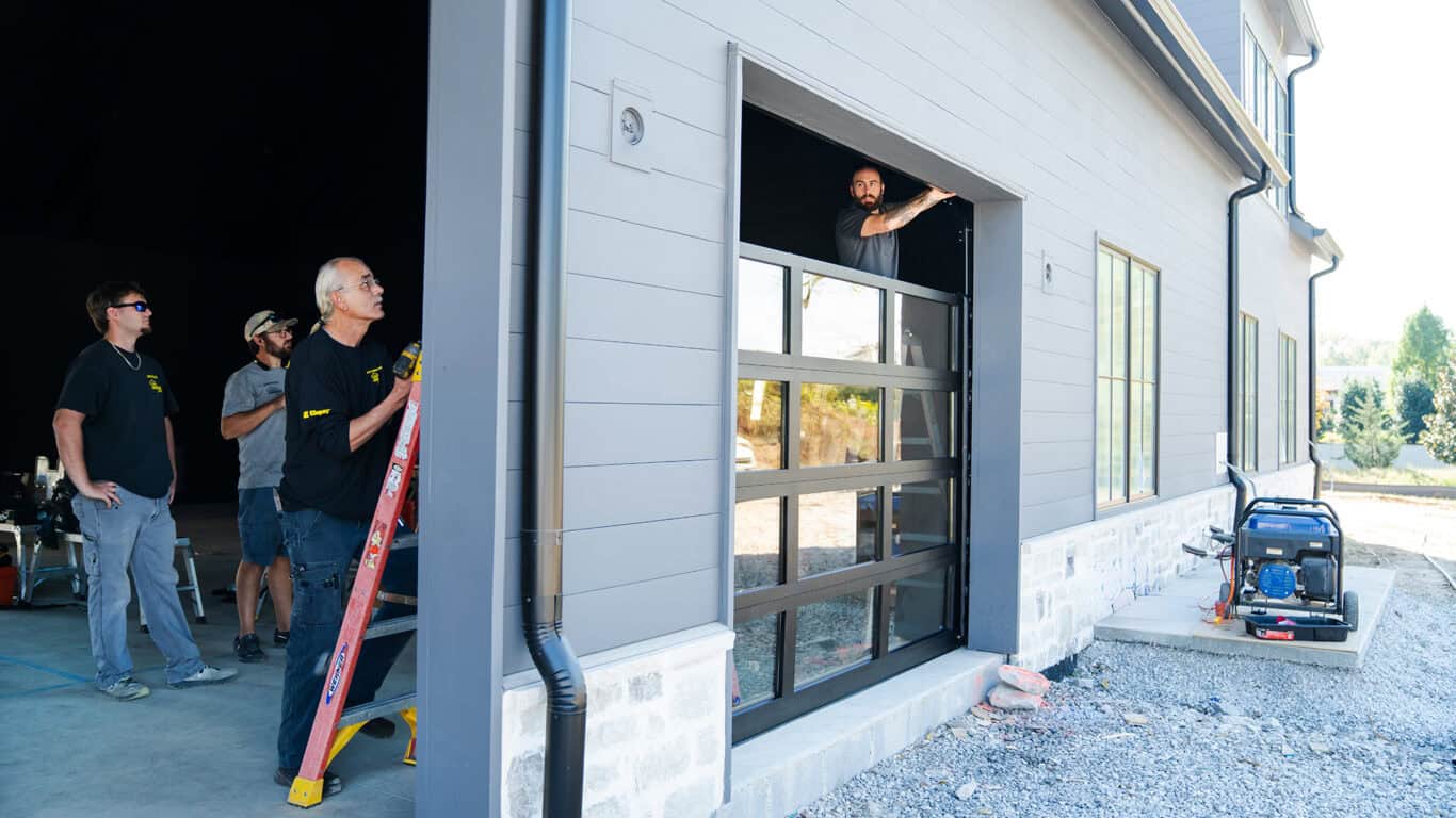 Workers are busy with a garage door installation on a modern building. One man stands on a ladder while others assist nearby, ensuring everything goes smoothly. A generator hums softly on the ground, ready for any emergency garage door service needs that might arise.