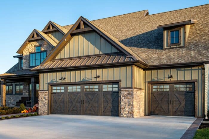 Large stone and wood house with three garage doors, gabled roof, and decorative elements. Lawn and walkway are visible in front.
