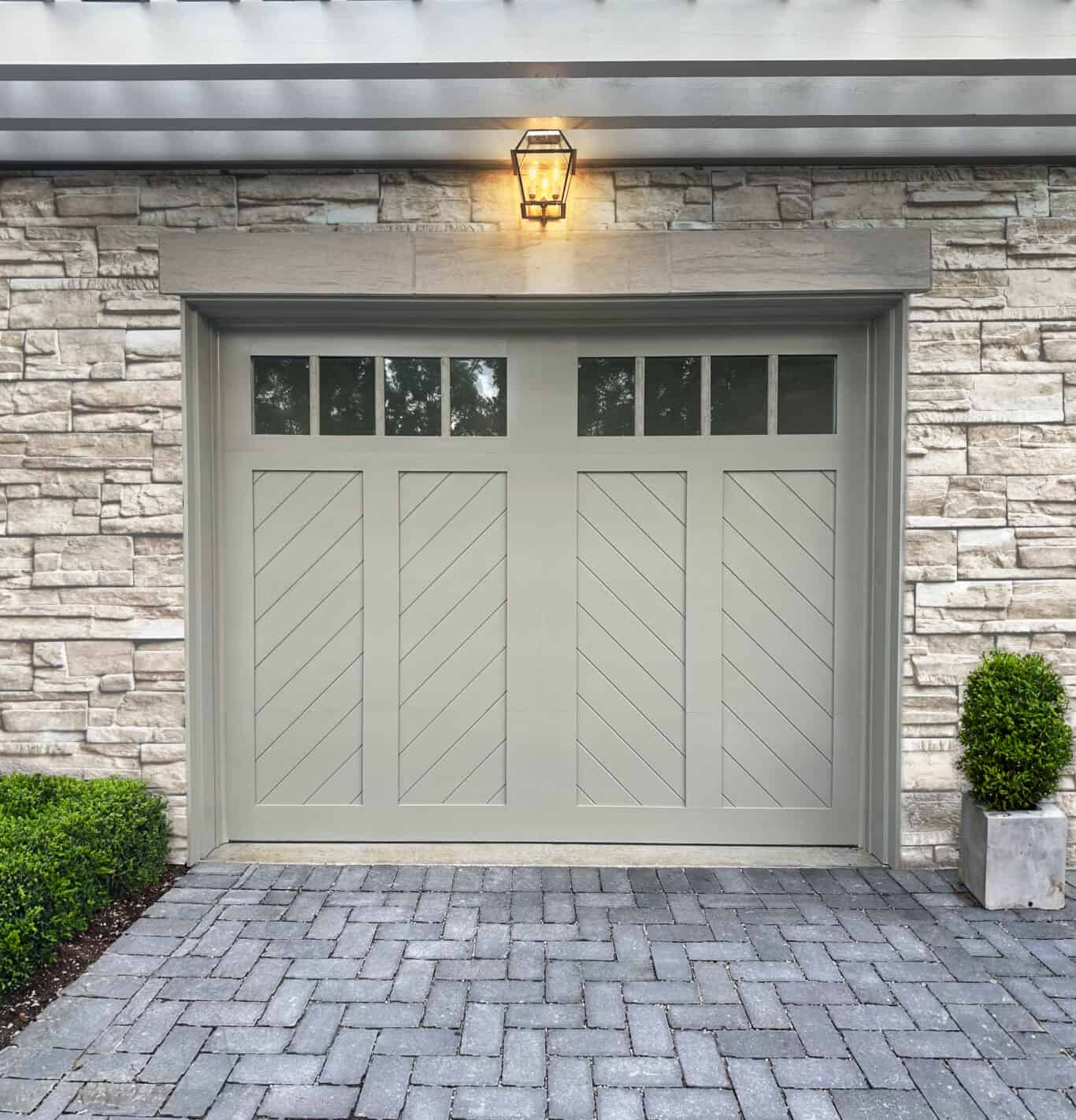 A residential garage door with diagonal panels and small windows at the top is set in a stone wall, complemented by a lantern above and potted plants on either side.