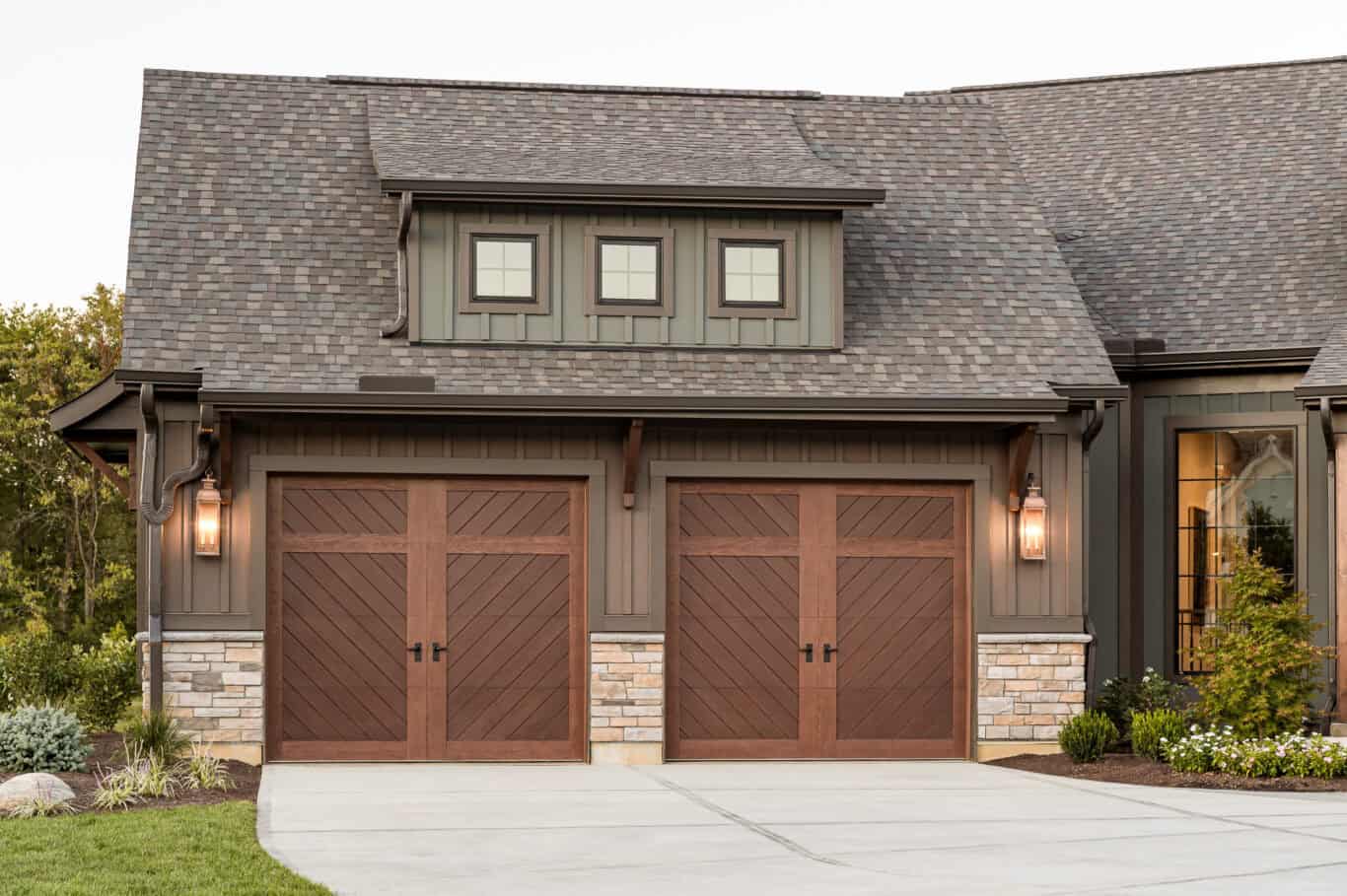 This residence features a two-car garage with residential garage doors, charming wooden doors, stone accents, and a shingle roof. Flanking the entrance are two elegant lantern-style lights that enhance its welcoming aura.
